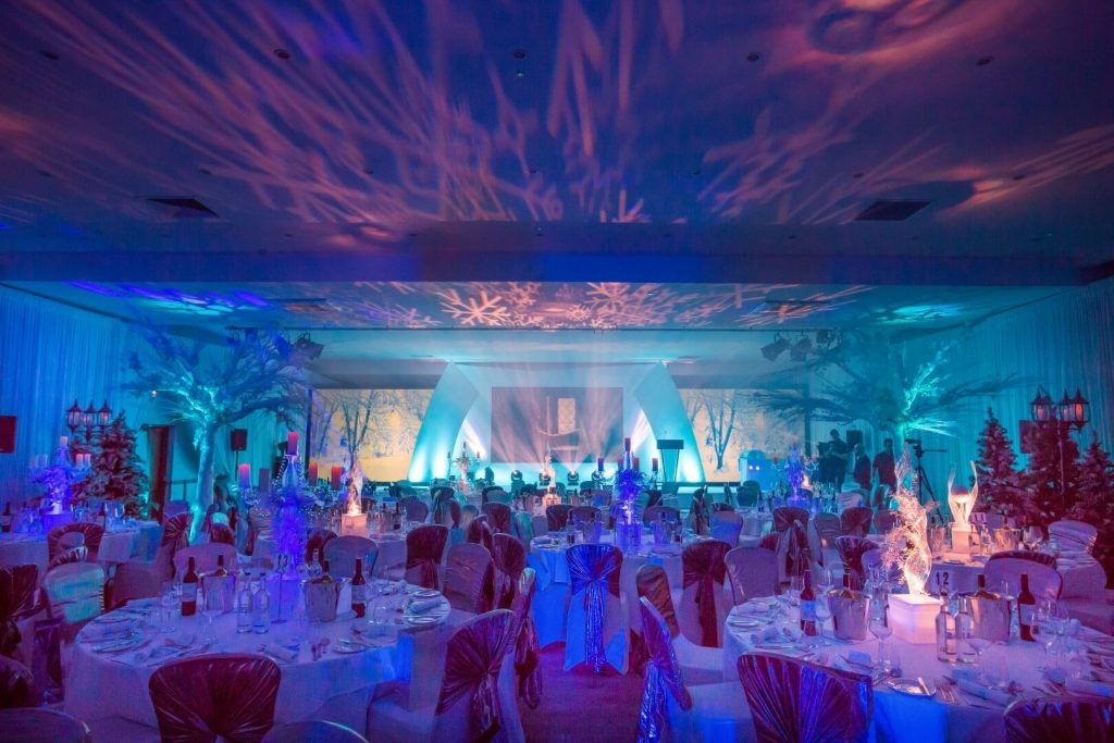 Narnia Winter Wonderland themed event snowflake av lighting on the ceiling blue uplighting on white artificial trees led transparent frosted table centres white table and chairs with gold shimmer sashes