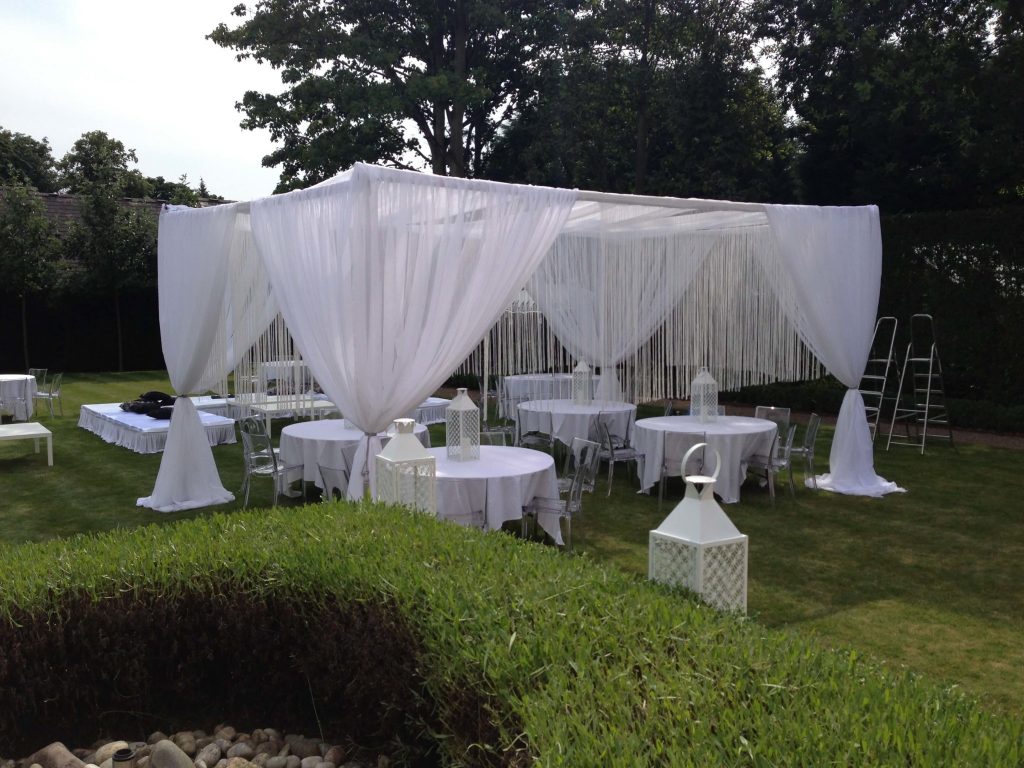 outdoor Summer event with white string curtain draping from a cabana with white tables underneath with white storm lanterns and transparent chivari chairs